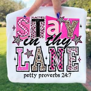 Peut inclure: T-shirt blanc avec l'inscription "Stay in thy Lane" en rose, noir et imprimé léopard. L'expression "petty proverbs 24:7" est imprimée en bas. Le t-shirt est tenu par une main.