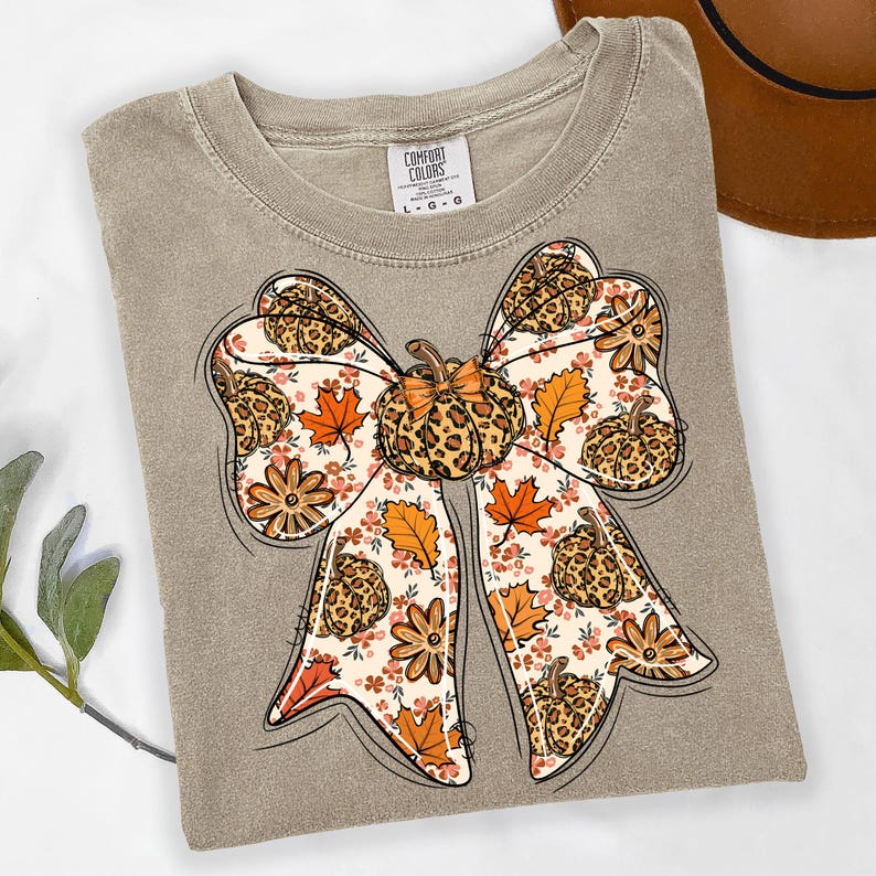 May include: A tan Comfort Colors t-shirt featuring a large bow design. The bow is decorated with a pumpkin, fall leaves, and floral patterns. The pumpkin and bow details have a leopard print design. The shirt is a size L-G-G.