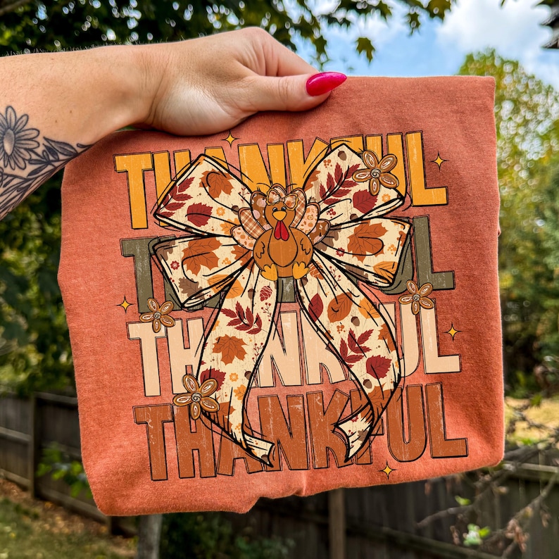 May include: A rust-colored t-shirt featuring the word "THANKFUL" in a layered design. A large bow with fall leaves and a turkey graphic is centered on the shirt. The shirt is held up by a hand with a floral tattoo.