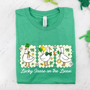 Peut inclure: T-shirt vert avec un motif de trois oies de dessin anim&eacute;, chacune avec des accessoires sur le th&egrave;me de la Saint-Patrick. Le texte "Lucky Goose on the Loose" est imprim&eacute; en dessous. Le t-shirt est pli&eacute;.