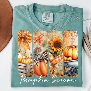 May include: A teal Comfort Colors t-shirt with a fall-themed graphic. The graphic includes a bow, flowers, a sunflower, and pumpkins. The words "Pumpkin Season" are written in a script font across the bottom of the graphic.