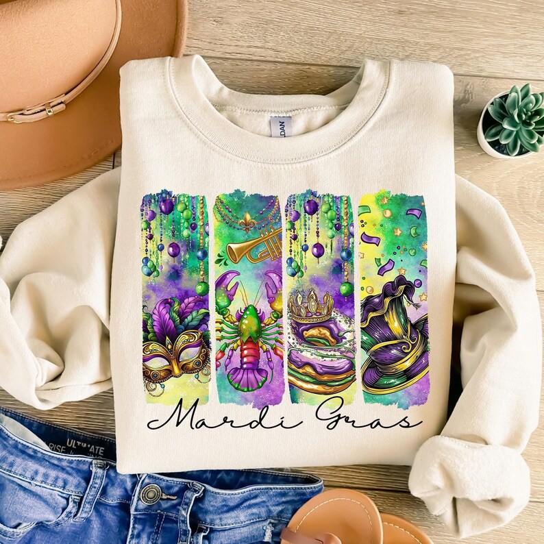 May include: A cream colored sweatshirt with a watercolor design of Mardi Gras symbols. The symbols include a mask, a lobster, a crown, and a hat. The text "Mardi Gras" is written in black below the symbols.