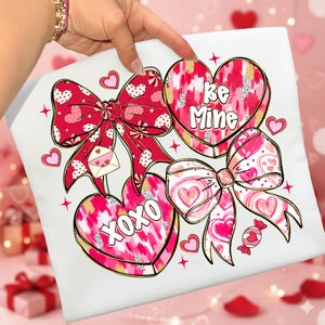 Puede incluir: Coj&iacute;n blanco con un dise&ntilde;o de San Valent&iacute;n. El dise&ntilde;o presenta lazos rojos, corazones y las frases "Be Mine" y "XOXO". Los lazos tienen patrones de corazones y los corazones tienen detalles dorados. Tambi&eacute;n se incluye un peque&ntilde;o sobre con un coraz&oacute;n.