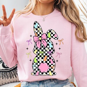 May include: Pink sweatshirt with a checkered bunny silhouette. The bunny is decorated with pastel-colored jelly beans, bows, and flowers.