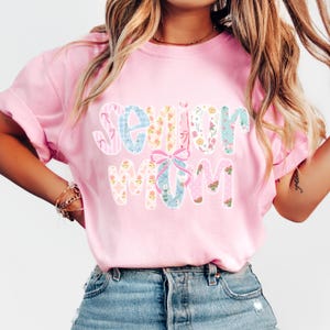 Puede incluir: Una camiseta rosa claro con la frase "Senior Mom" en un dise&ntilde;o floral colorido. Las letras presentan varios patrones, incluyendo rayas, flores y lunares. Un lazo rosa se encuentra entre las palabras.