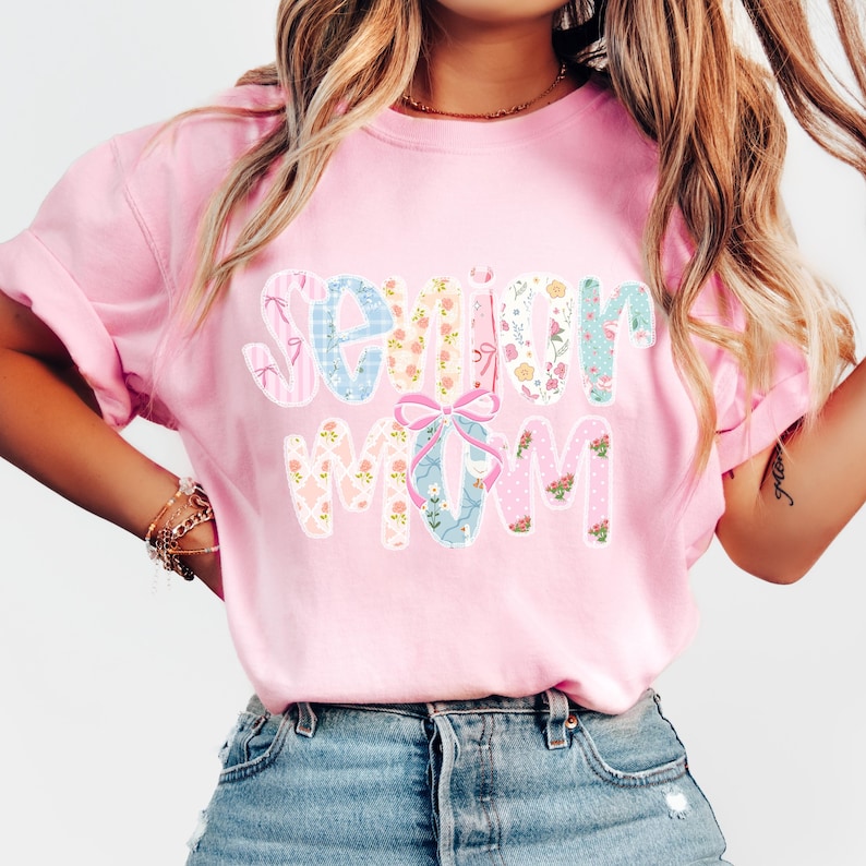 Puede incluir: Una camiseta rosa claro con la frase "Senior Mom" en un dise&ntilde;o floral colorido. Las letras presentan varios patrones, incluyendo rayas, flores y lunares. Un lazo rosa se encuentra entre las palabras.