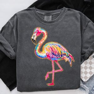 May include: A dark gray sweatshirt featuring a colorful flamingo design. The flamingo is depicted with vibrant rainbow hues, outlined in gold glitter. The sweatshirt has a crew neck and long sleeves, with a relaxed fit.