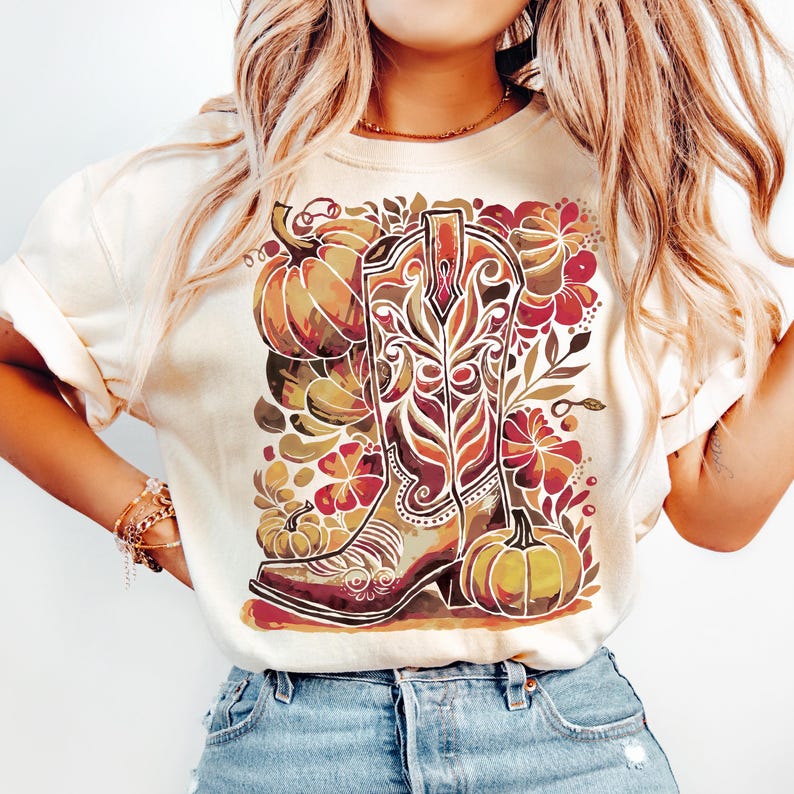 Op de afbeelding: Cr&egrave;mekleurig T-shirt met een gedetailleerde illustratie van een cowboylaars omringd door pompoenen en bloemelementen in bruin-, oranje- en roodtinten. Het ontwerp heeft een aquareleffect, waardoor een rustieke en herfstachtige esthetiek ontstaat.
