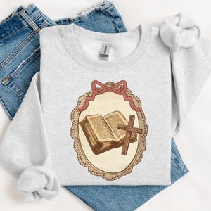 May include: A light gray sweatshirt featuring a detailed illustration of an open book and a wooden cross within a decorative oval frame. The frame is adorned with a pink bow. The sweatshirt is laid on top of blue jeans.