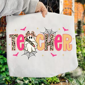 Pu&ograve; includere: Borsa tote bianca con la scritta "TEACHER" in lettere rosa e beige con motivo leopardato. Un fantasma con un fiocco e una matita &egrave; al centro. Pipistrelli rosa e ragnatele decorano il disegno.
