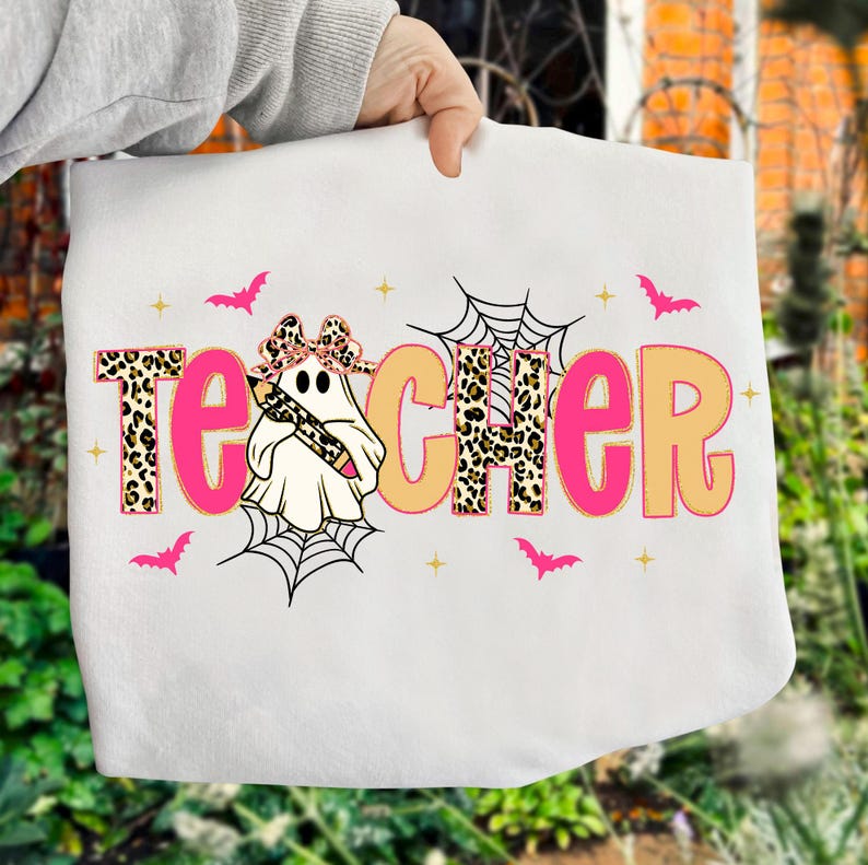 Pu&ograve; includere: Borsa tote bianca con la scritta "TEACHER" in lettere rosa e beige con motivo leopardato. Un fantasma con un fiocco e una matita &egrave; al centro. Pipistrelli rosa e ragnatele decorano il disegno.
