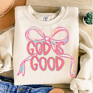 May include: Cream-colored sweatshirt with a large, colorful bow design and the words "GOD IS GOOD" in pink. The bow features pastel rainbow stripes. The sweatshirt is a casual, comfortable style.