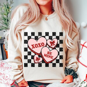 May include: A cream-colored sweatshirt with a checkered pattern and three pink candy hearts with red glitter. The hearts say "XOXO", "BE MINE", and have a bow.