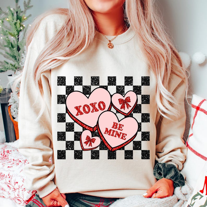 May include: A cream-colored sweatshirt with a checkered pattern and three pink candy hearts with red glitter. The hearts say "XOXO", "BE MINE", and have a bow.