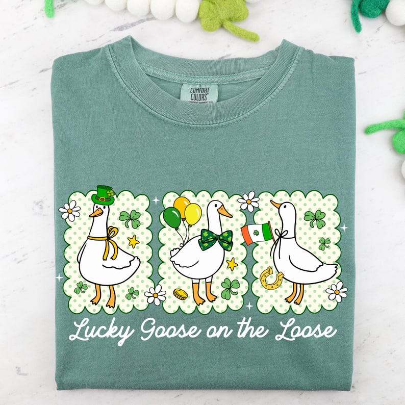 Peut inclure: T-shirt vert avec un motif de trois oies de dessin anim&eacute;, chacune avec des accessoires sur le th&egrave;me de la Saint-Patrick et irlandais. Le texte "Lucky Goose on the Loose" est imprim&eacute; en dessous.