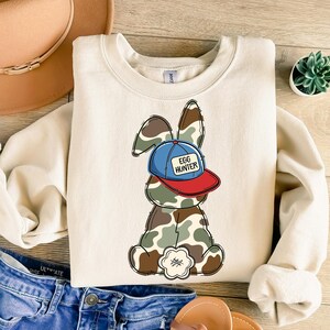 May include: A cream-colored sweatshirt with a cartoon bunny wearing a blue and red baseball cap that says "Egg Hunter". The bunny is wearing camouflage.