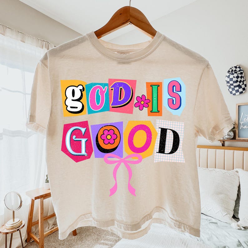May include: A beige t-shirt with the words "GOD IS GOOD" in colorful, cut-out style letters. The design includes a pink bow and flower accents. The shirt is hanging on a wooden hanger.