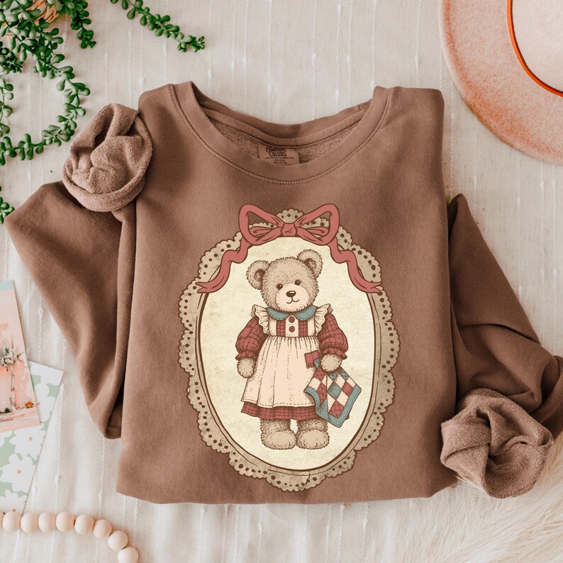 Op de afbeelding: Een bruine sweatshirt met een vintage illustratie van een teddybeer. De beer draagt een jurk en houdt een quilt vast, omlijst door een ovaal met een roze strik. De sweatshirt heeft lange mouwen.