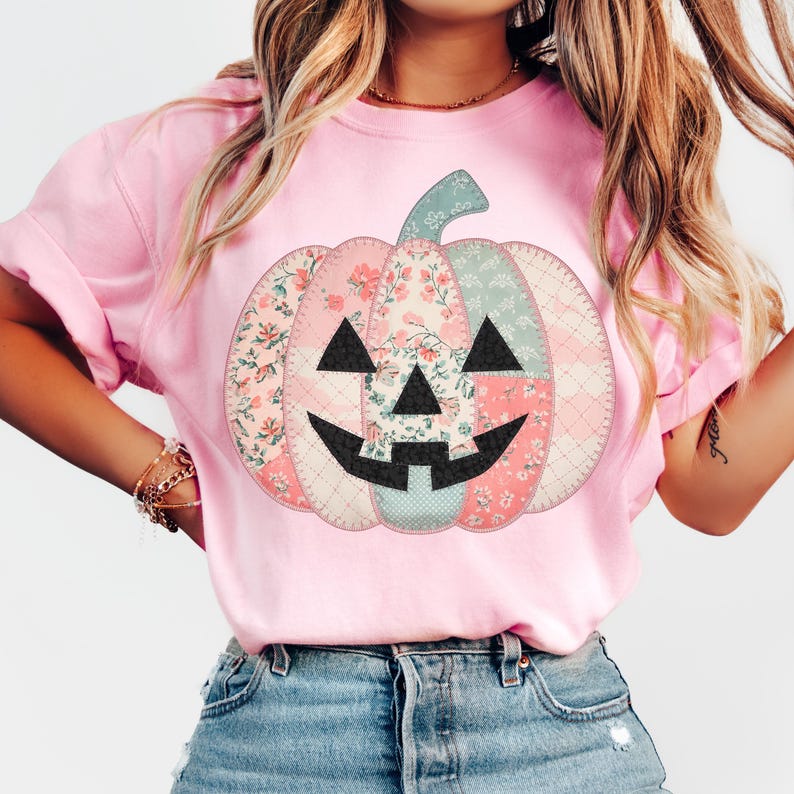 May include: A light pink t-shirt featuring a patchwork pumpkin design. The pumpkin has a green stem and is composed of floral and patterned fabric in pink, blue, and white. The pumpkin's face is black.