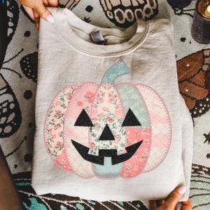 May include: A light beige sweatshirt featuring a patchwork pumpkin design. The pumpkin is composed of floral and patterned fabrics in pink, blue, and cream, with black triangle eyes and a smiling mouth. The stem is light blue.