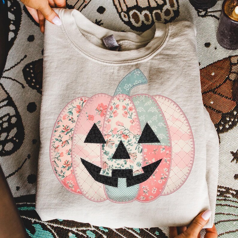 May include: A light beige sweatshirt featuring a patchwork pumpkin design. The pumpkin is composed of floral and patterned fabrics in pink, blue, and cream, with black triangle eyes and a smiling mouth. The stem is light blue.