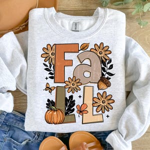 May include: A light gray sweatshirt with the word "FALL" in brown and tan block letters. The design includes fall-themed graphics such as flowers, a pumpkin, an acorn, a leaf, and a butterfly. The sweatshirt is folded on top of blue jeans and sandals.