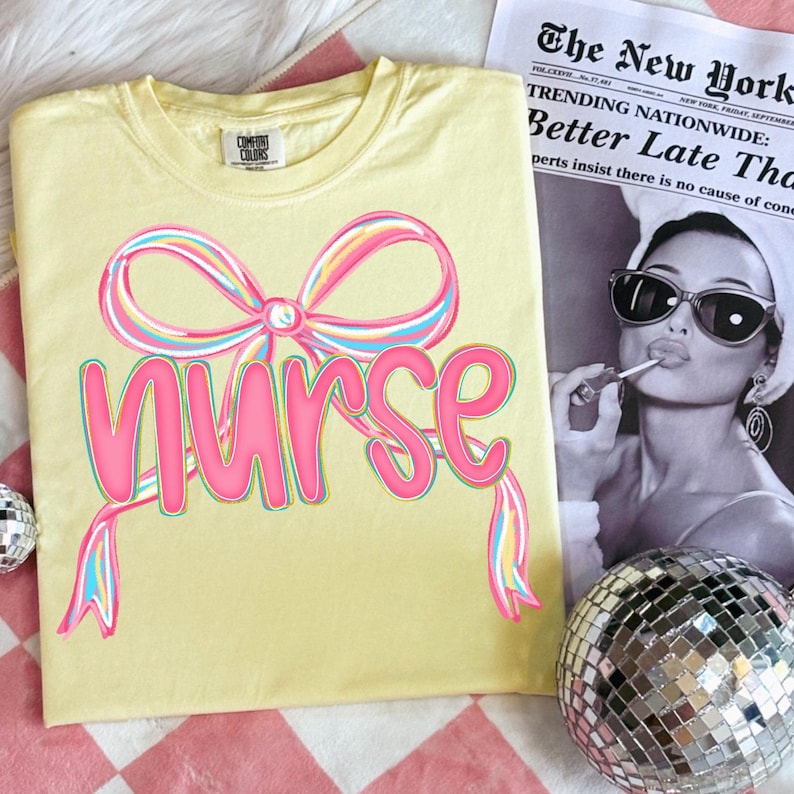 Pode incluir: Camiseta amarelo claro com a palavra "Nurse" em rosa, adornada com um design de la&ccedil;o colorido. A camiseta &eacute; exibida com um jornal e uma bola de discoteca.