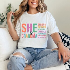 May include: White t-shirt with the text "SHE is GRANDMA" in rainbow colors. The text is stacked vertically with the words "STRONG, CHOSEN, BEAUTIFUL, CAPABLE, VICTORIOUS, NEVER ALONE, GREATLY LOVED" stacked below in smaller font.