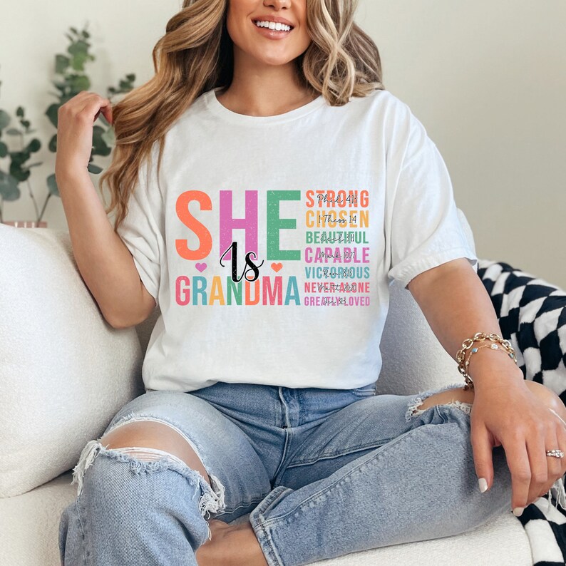 May include: White t-shirt with the text "SHE is GRANDMA" in rainbow colors. The text is stacked vertically with the words "STRONG, CHOSEN, BEAUTIFUL, CAPABLE, VICTORIOUS, NEVER ALONE, GREATLY LOVED" stacked below in smaller font.