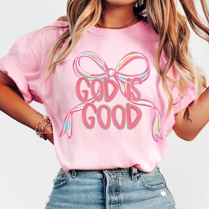 May include: A pink t-shirt with the words "GOD IS GOOD" in a colorful, outlined font. The text is centered below a large bow with rainbow-colored accents. The shirt is a solid pink color.