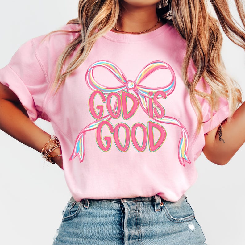 May include: A pink t-shirt with the words "GOD IS GOOD" in a colorful, outlined font. The text is centered below a large bow with rainbow-colored accents. The shirt is a solid pink color.