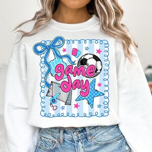 May include: White sweatshirt with a colorful soccer-themed graphic. The design includes a soccer ball, stars, a bow, and the words "Game Day" in pink and blue. The sweatshirt has a crew neck and long sleeves.