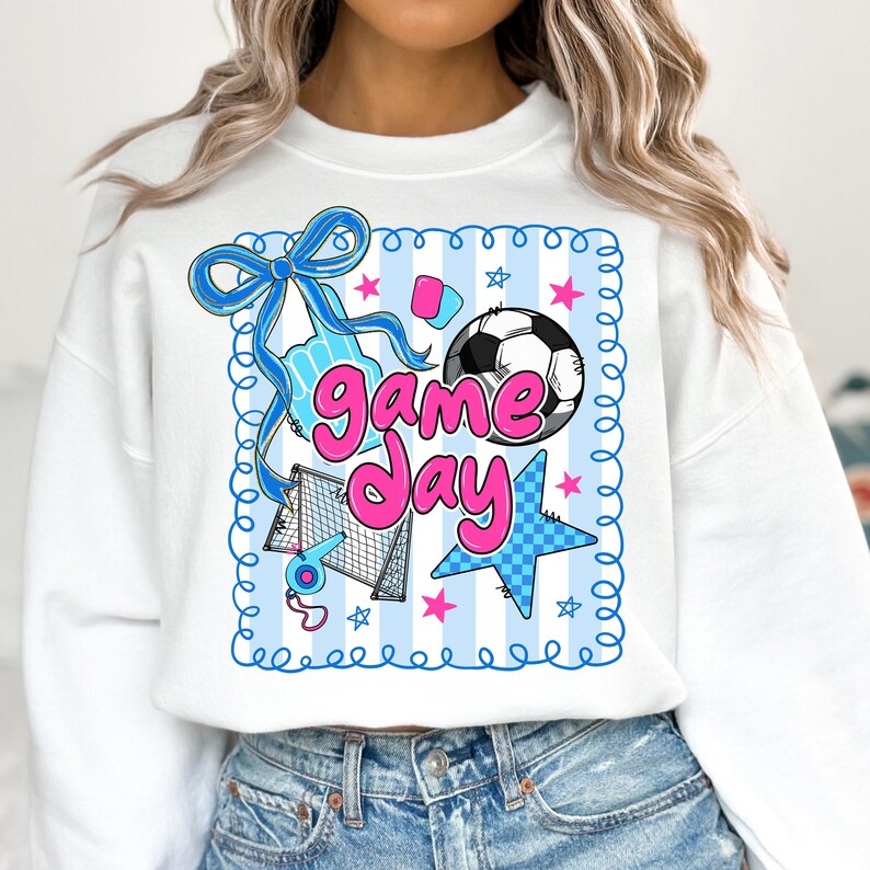 May include: White sweatshirt with a colorful soccer-themed graphic. The design includes a soccer ball, stars, a bow, and the words "Game Day" in pink and blue. The sweatshirt has a crew neck and long sleeves.