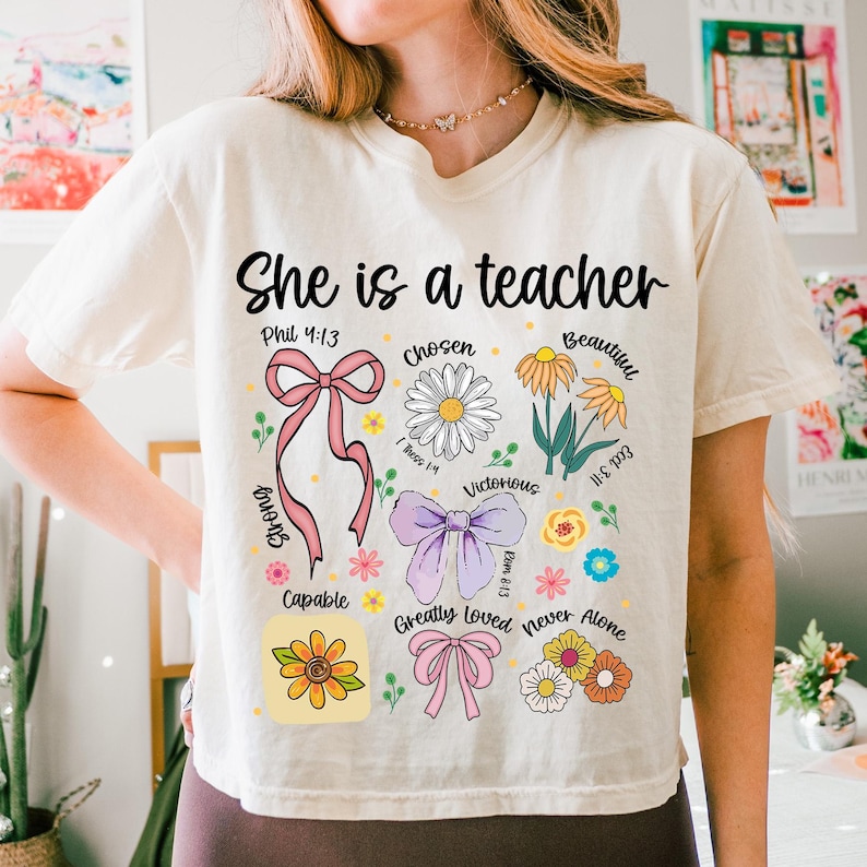 Op de afbeelding: Cr&egrave;mekleurig T-shirt met de tekst "She is a teacher" in zwart schrift. Het shirt heeft bloemen- en lintafbeeldingen met woorden als "Chosen", "Beautiful" en "Capable".