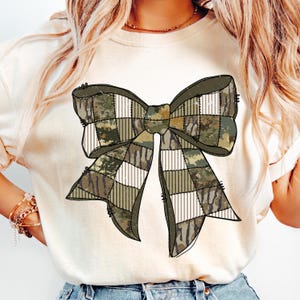 May include: A cream-colored t-shirt featuring a large bow design. The bow is composed of various camouflage and plaid patterns in shades of green, brown, and white. The bow is centered on the shirt, creating a rustic and stylish look.