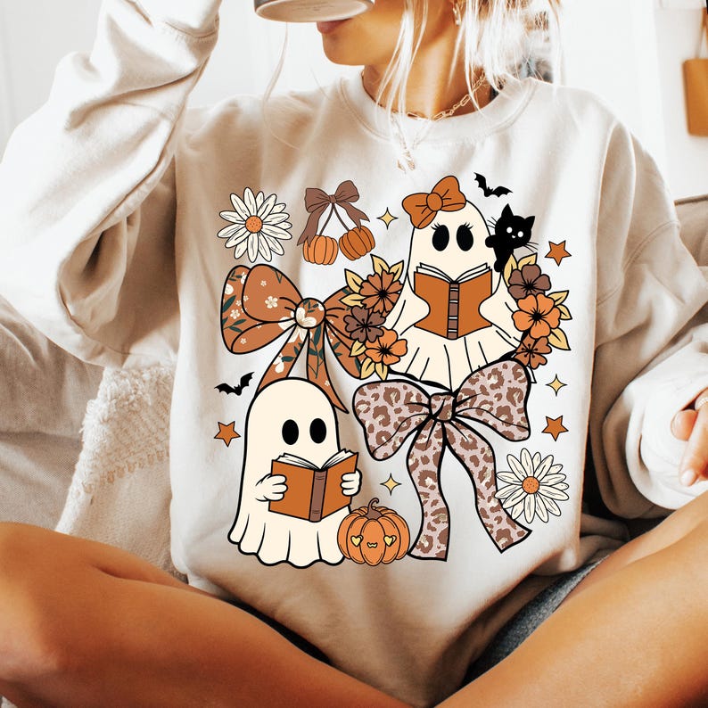 Op de afbeelding: Cr&egrave;mekleurige sweatshirt met een grillig Halloween-ontwerp. De afbeelding bevat twee spookkarakters die boeken lezen, omringd door bloemstukken, strikken, pompoenen, sterren en een zwarte kat. Het ontwerp bevat tinten bruin, oranje en cr&egrave;me.
