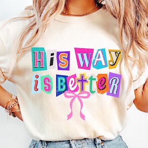 May include: Cream-colored sweatshirt with the text "His Way is Better" in colorful, cut-out style letters. A pink bow is below the text. The sweatshirt is a casual, comfortable style.
