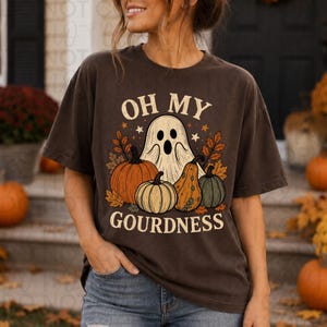 May include: A dark brown short-sleeve t-shirt featuring a graphic print of a white ghost surrounded by various pumpkins and autumn leaves. The text "OH MY GOURDNESS" is printed in a cream color above and below the central illustration.
