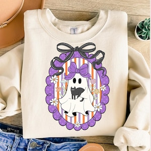 May include: Cream-colored sweatshirt featuring a Halloween-themed graphic. The design includes a cartoon ghost holding a black cat, framed by a purple oval with flowers and a black bow. The background has orange and white stripes.