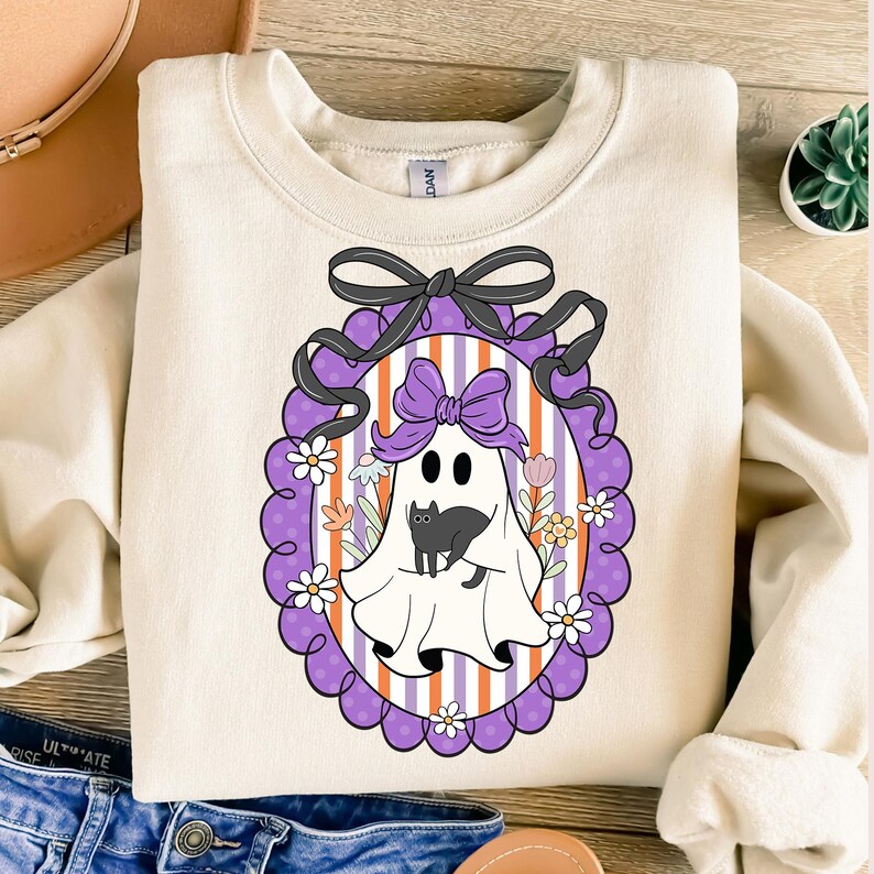 May include: Cream-colored sweatshirt featuring a Halloween-themed graphic. The design includes a cartoon ghost holding a black cat, framed by a purple oval with flowers and a black bow. The background has orange and white stripes.