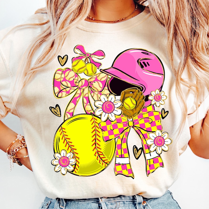 May include: A cream-colored t-shirt featuring a softball-themed graphic. The design includes a yellow softball, a pink helmet, a brown glove, and decorative bows with a pink and yellow checkered pattern. Floral accents and heart shapes complete the design.