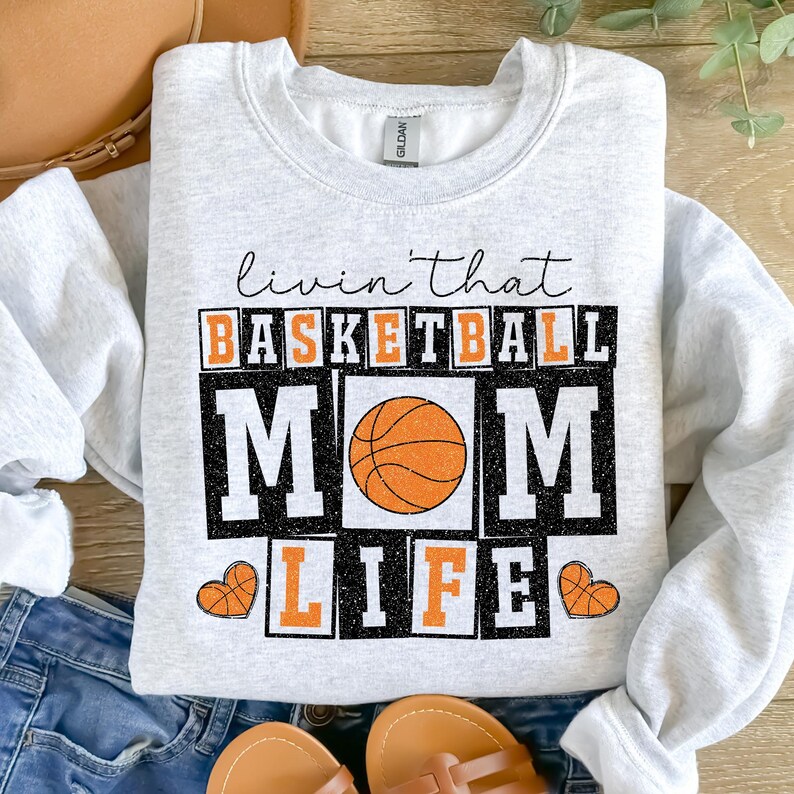 May include: A gray sweatshirt with the text "Livin' that Basketball Mom Life" in black and white with a basketball graphic. The text is in a blocky font with a glittery effect.