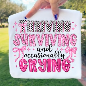 以下が含まれることがあります： 白のスウェットシャツに「Thriving, Surviving, and occasionally Crying」の文字が黒とピンクでプリントされています。「Thriving」の文字は白黒のチェック柄です。「Surviving」の文字はピンクのヒョウ柄です。