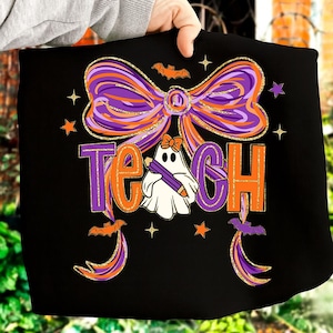May include: Black t-shirt with a Halloween-themed design. The design features the word "TEACH" in orange and purple, a ghost holding a pencil, a large bow, bats, and stars. The bow and text have a gold glitter effect.