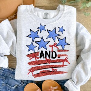 Pode incluir: Uma camisa de moletom cinza com um design de bandeira americana vermelha, branca e azul. A bandeira &eacute; feita de glitter e tem estrelas e la&ccedil;os vermelhos. A palavra "AND" est&aacute; escrita em glitter preto sobre a bandeira.