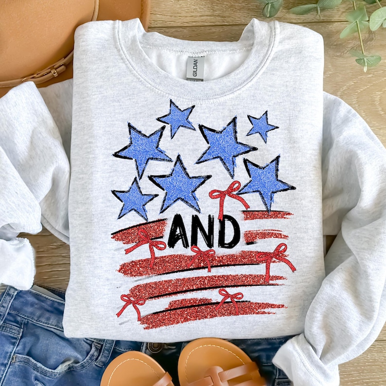 Pode incluir: Uma camisa de moletom cinza com um design de bandeira americana vermelha, branca e azul. A bandeira &eacute; feita de glitter e tem estrelas e la&ccedil;os vermelhos. A palavra "AND" est&aacute; escrita em glitter preto sobre a bandeira.