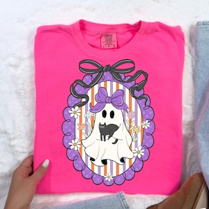 May include: A bright pink Comfort Colors sweatshirt featuring a Halloween-themed graphic. The graphic includes a white ghost with a purple bow, a black cat, and a decorative oval frame with flowers and a black bow.