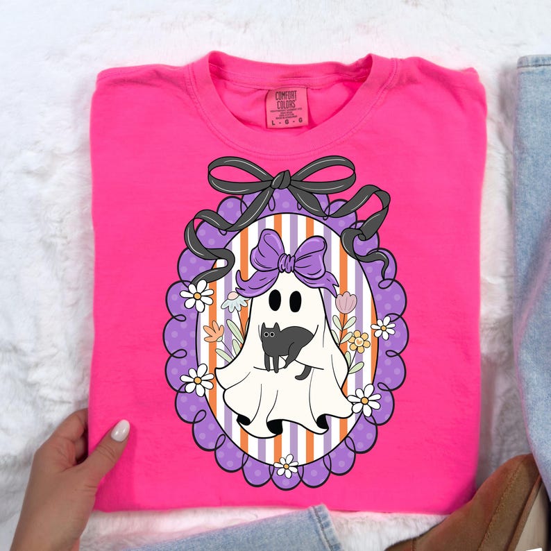 May include: A bright pink Comfort Colors sweatshirt featuring a Halloween-themed graphic. The graphic includes a white ghost with a purple bow, a black cat, and a decorative oval frame with flowers and a black bow.