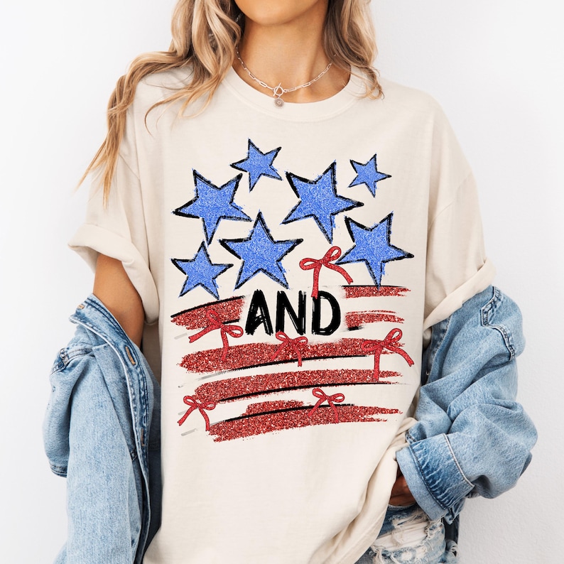 Pode incluir: Uma camiseta branca com um design de bandeira americana vermelha, branca e azul. A bandeira &eacute; feita de estrelas e listras com la&ccedil;os vermelhos. A palavra "AND" est&aacute; escrita em glitter vermelho na bandeira.