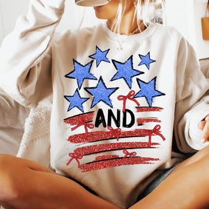 Pode incluir: Camisola branca com um design de bandeira americana vermelha, branca e azul. A bandeira tem listras vermelhas, estrelas brancas e la&ccedil;os vermelhos. A palavra "AND" est&aacute; escrita em letras pretas na bandeira.